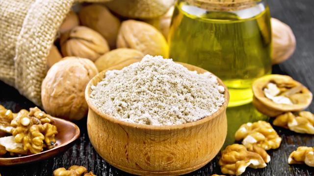 Walnut flour in bowl, nuts on a table, in spoon and in a jute bag, butter in glass jar on background of dark wooden board