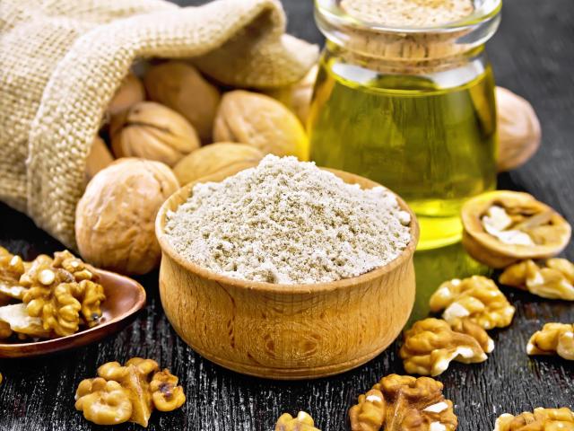 Walnut flour in bowl, nuts on a table, in spoon and in a jute bag, butter in glass jar on background of dark wooden board