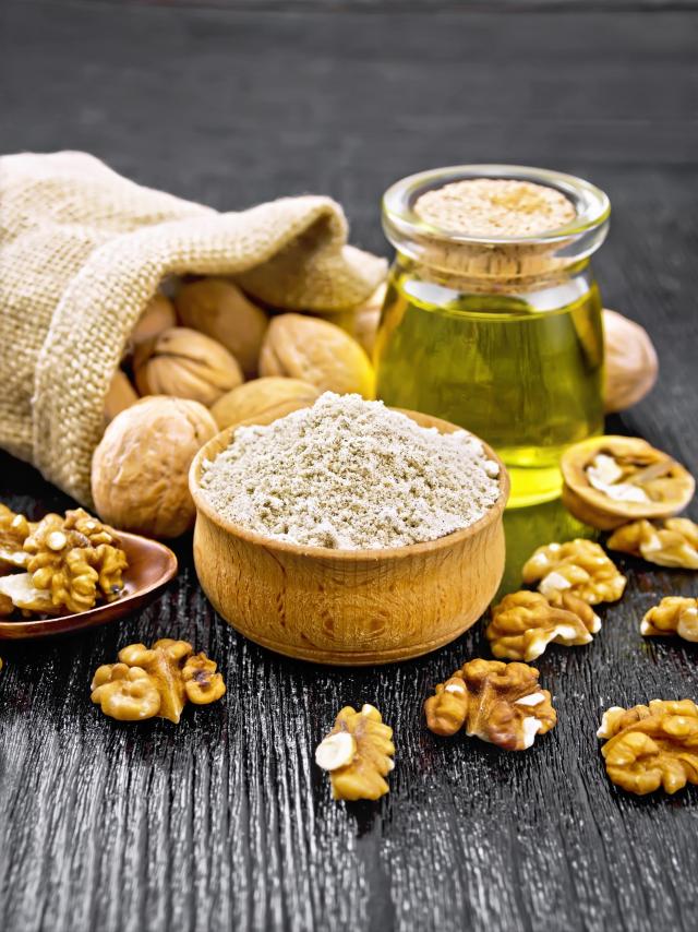 Walnut flour in bowl, nuts on a table, in spoon and in a jute bag, butter in glass jar on background of dark wooden board